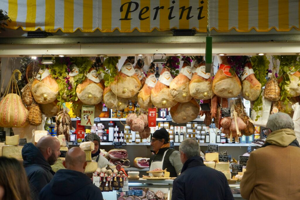 Authentic Italian market stall Perini displaying hanging prosciutto, aged cheeses, and gourmet products in Florence - traditional food markets and culinary shopping experiences in Tuscany for epicurean travelers